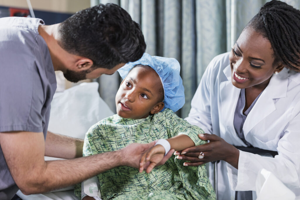 Uma menina afro-americana de 6 anos sentada numa maca do hospital, com um médico e enfermeiro a cuidar dela.  Está a usar uma bata hospitalar e touca cirúrgica. Um enfermeiro de bata, um jovem médio-oriental na casa dos 20 anos, está de pé de um dos lados dela, e uma médica, uma mulher afro-americana madura nos seus 40 anos, está do outro lado.