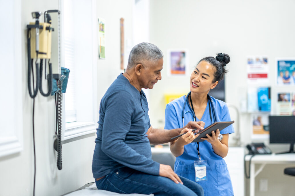 Um cavalheiro sénior de descendência hispânica senta-se numa mesa de exame durante um exame de rotina nos médicos.  Um enfermeiro, que está vestido de uniforme azul, está de pé em frente ao cavalheiro enquanto analisa alguns resultados recentes de testes com a doente num tablet.